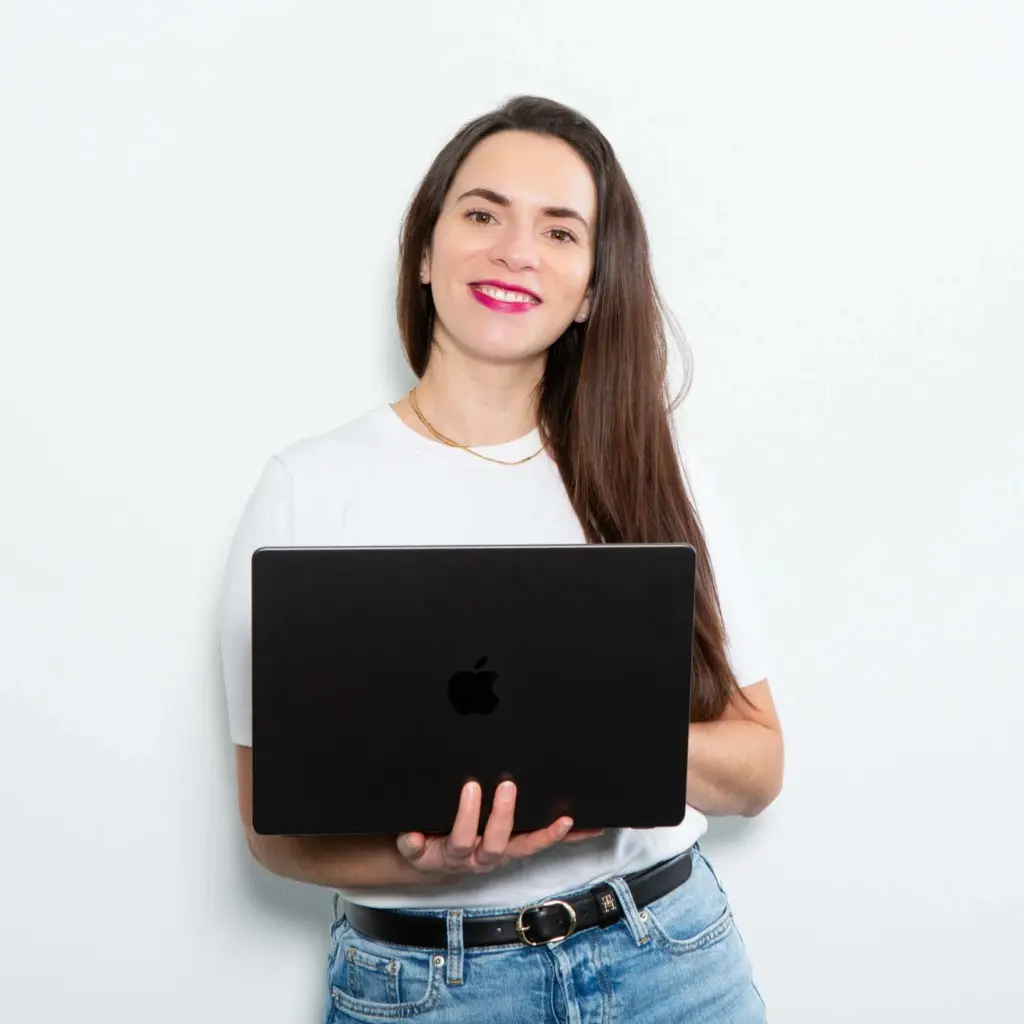 Eine lächelnde Frau mit langen braunen Haaren hält einen schwarzen Laptop in den Händen und steht vor einer weißen Wand.